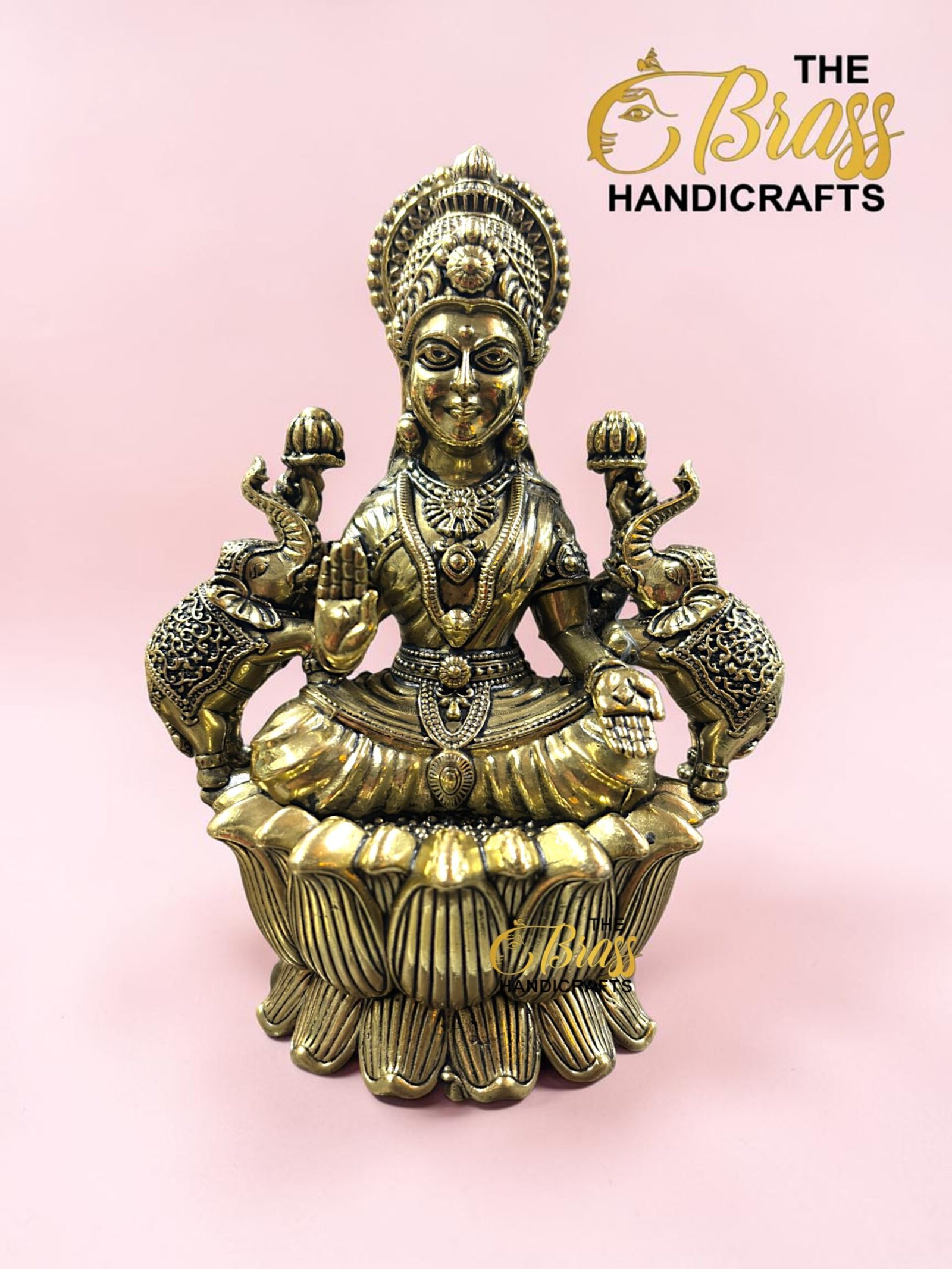 IMG_5773 - thebrasshandicrafts.com Golden brass statue of a Hindu goddess seated on a lotus pedestal with an ornate headdress, set against a pink background with a logo in the corner. thebrasshandicrafts.com