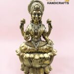 IMG_5774 - thebrasshandicrafts.com Gold-toned statue of a Hindu goddess seated on a lotus, ornate jewelry, against a pink background (The Brass Handicrafts). thebrasshandicrafts.com
