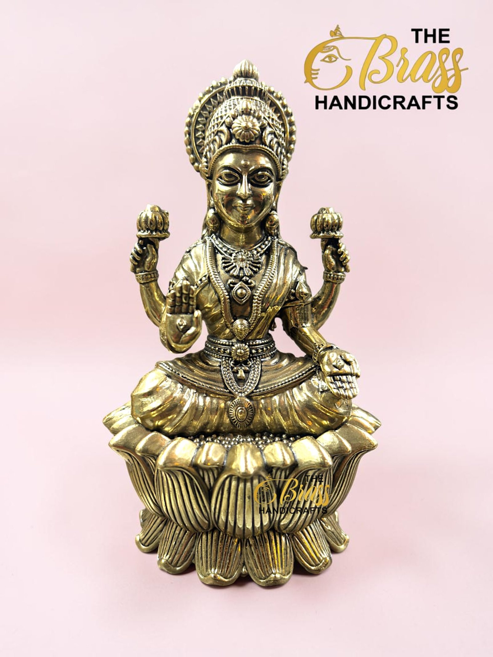 IMG_5774 - thebrasshandicrafts.com Gold-toned statue of a Hindu goddess seated on a lotus, ornate jewelry, against a pink background (The Brass Handicrafts). thebrasshandicrafts.com