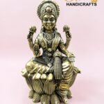 IMG_5776 - thebrasshandicrafts.com Brass statue of a Hindu goddess seated on a lotus with ornate headdress, against a pink background and logo reading 'The Brass Handicrafts'. thebrasshandicrafts.com