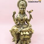 IMG_5778 - thebrasshandicrafts.com Gold statue of a seated Hindu goddess on a lotus, with an ornate crown and jewelry, against a pink background. thebrasshandicrafts.com