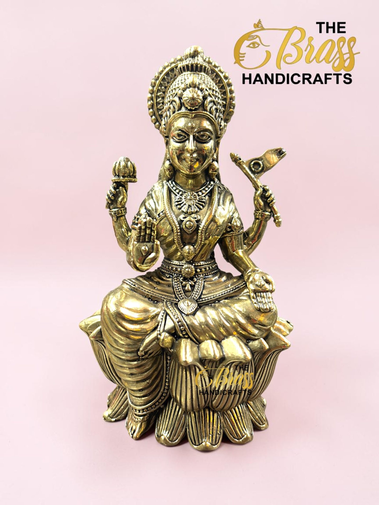 IMG_5778 - thebrasshandicrafts.com Gold statue of a seated Hindu goddess on a lotus, with an ornate crown and jewelry, against a pink background. thebrasshandicrafts.com