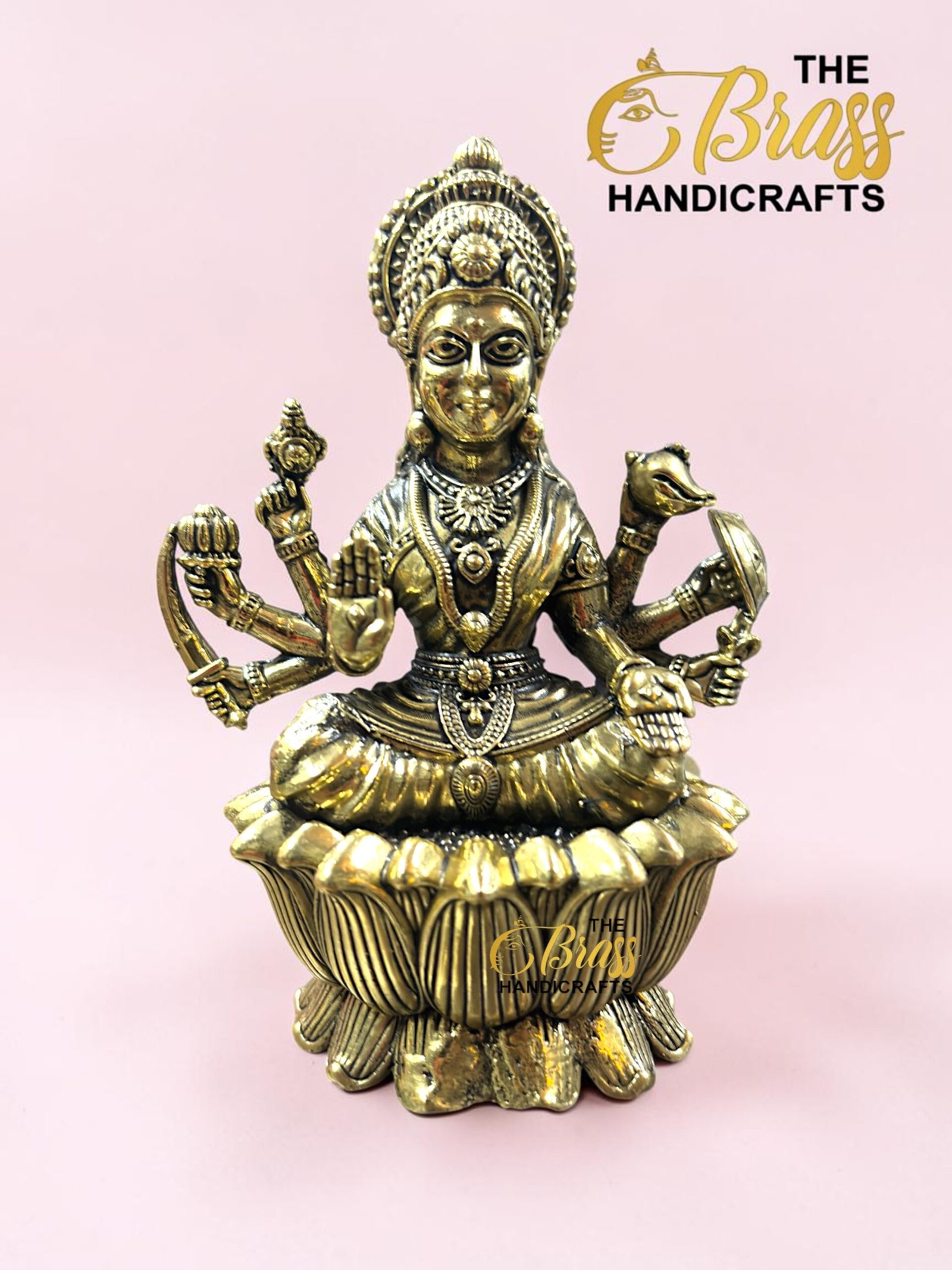 IMG_5779 - thebrasshandicrafts.com Gold-toned multi-armed deity statue with an ornate headdress, seated on a lotus pedestal against a pink background; branding for The Brass Handicrafts in the corner thebrasshandicrafts.com