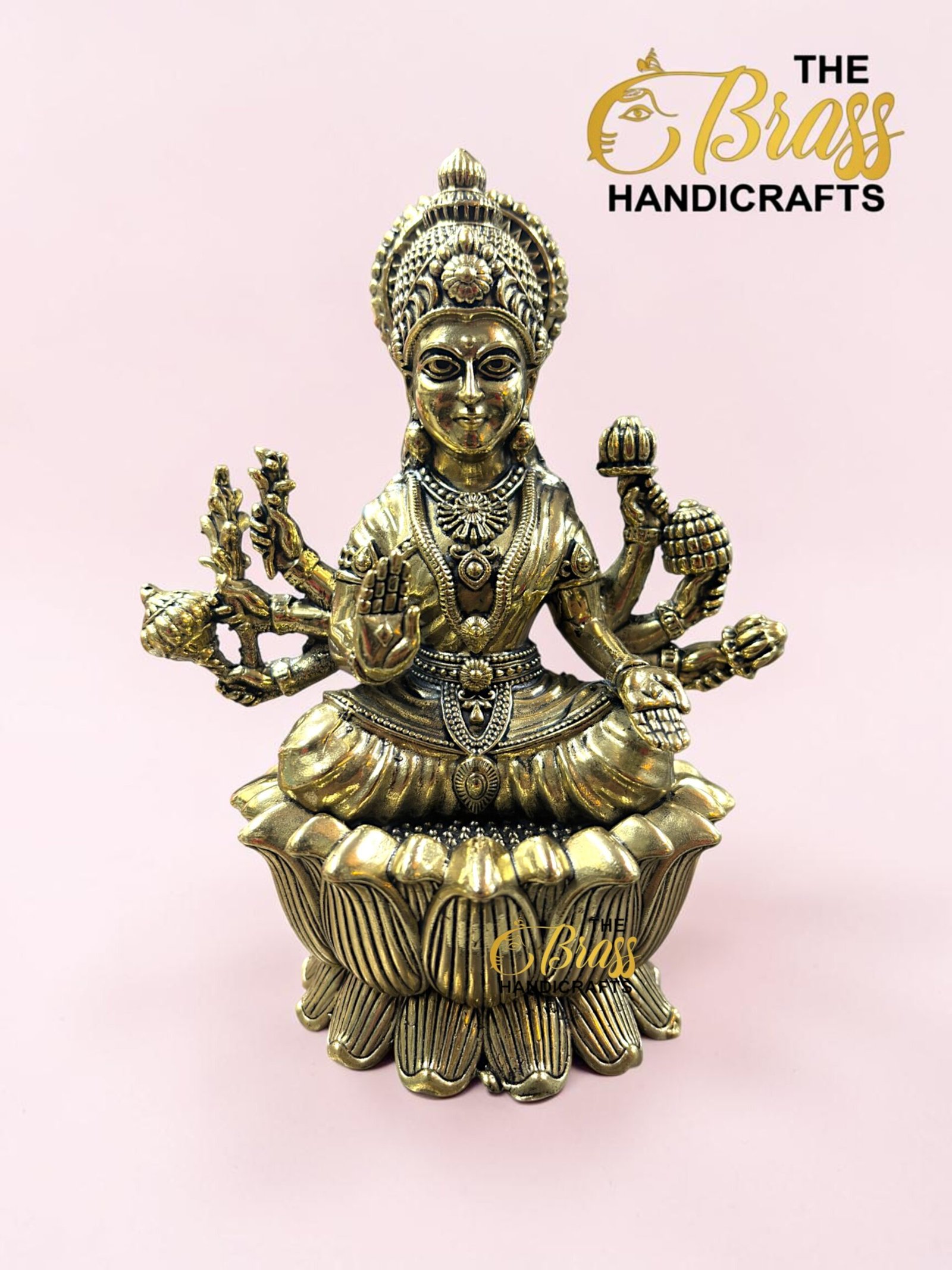 IMG_5780 - thebrasshandicrafts.com Gold brass deity statue with multiple arms and ornate headdress seated on a lotus base against a pink background; logo watermark reads 'The Brass Handicrafts'. thebrasshandicrafts.com