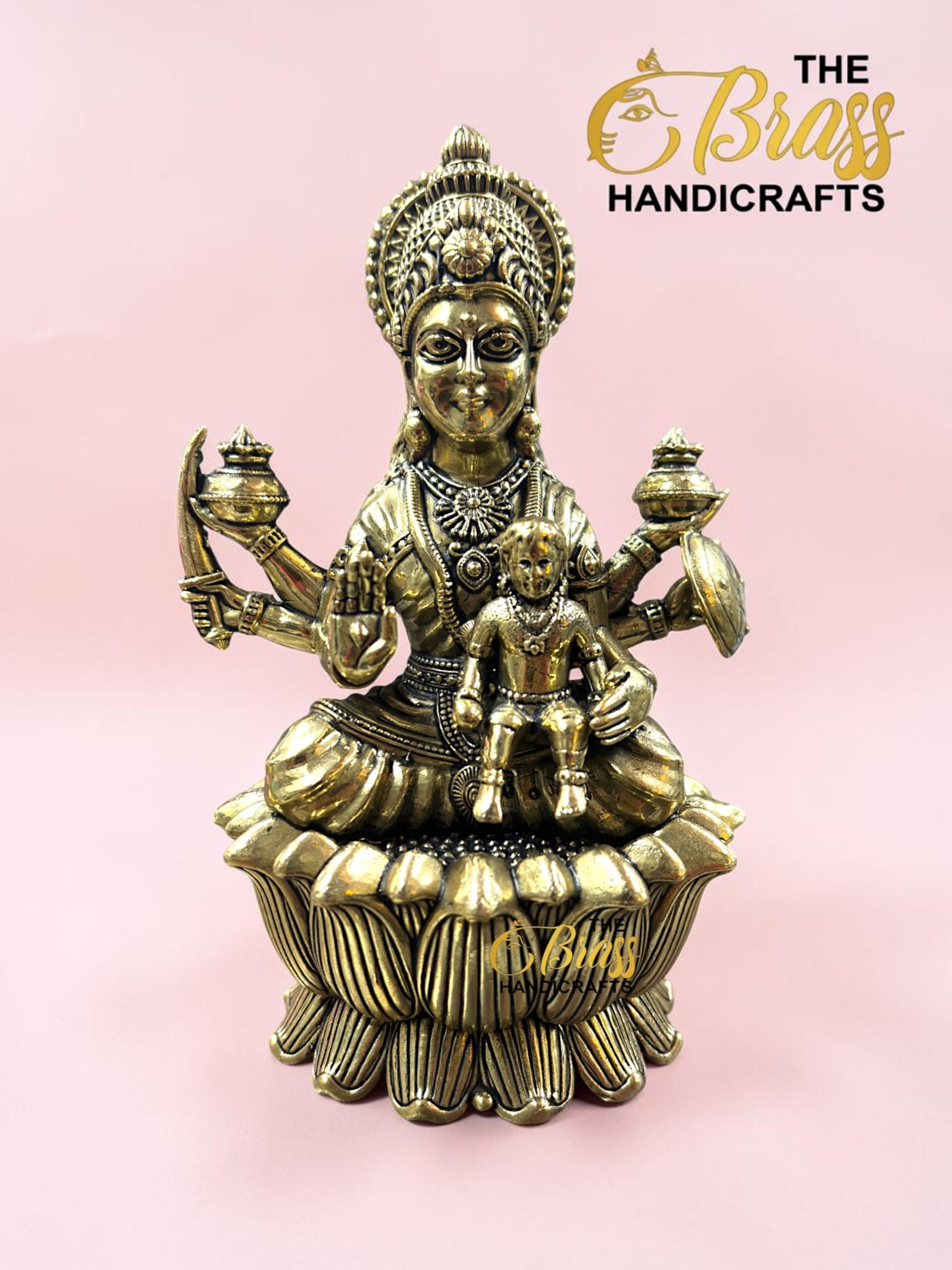 IMG_5781 - thebrasshandicrafts.com Brass goddess statue with child on a lotus base, pink background, logo reads The Brass Handicrafts. thebrasshandicrafts.com