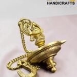 Close-up of an ornate gold pocket watch with a decorative chain on a pale pink surface.  thebrasshandicrafts.com