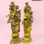IMG_5925 - thebrasshandicrafts.com Two ornate gold brass statues adorned with colorful stones on decorative bases against a pink backdrop; The Brass Handicrafts logo in the corner. thebrasshandicrafts.com