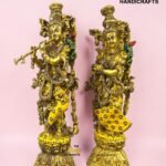 IMG_5926 - thebrasshandicrafts.com Two ornate gold brass deity statues with intricate jewelry and colorful stones, placed on decorative bases against a pink background. Watermark reads 'The Brass Handicrafts.' thebrasshandicrafts.com