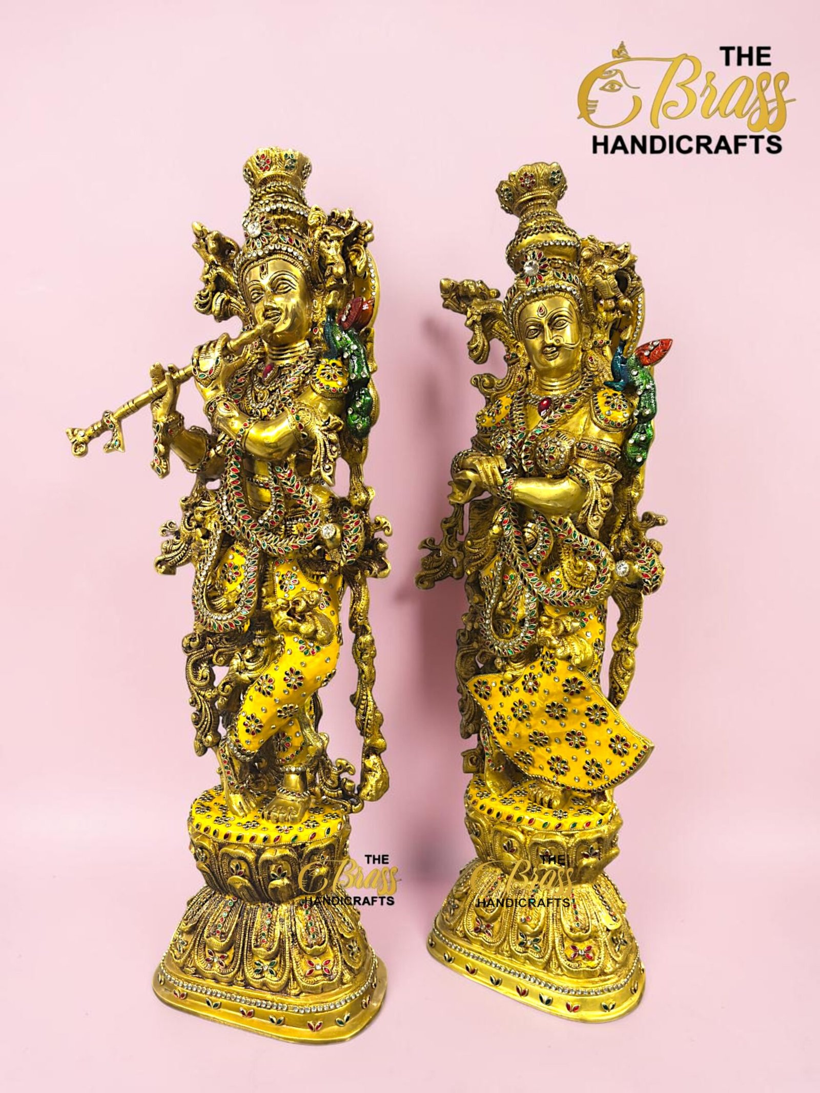 IMG_5926 - thebrasshandicrafts.com Two ornate gold brass deity statues with intricate jewelry and colorful stones, placed on decorative bases against a pink background. Watermark reads 'The Brass Handicrafts.' thebrasshandicrafts.com