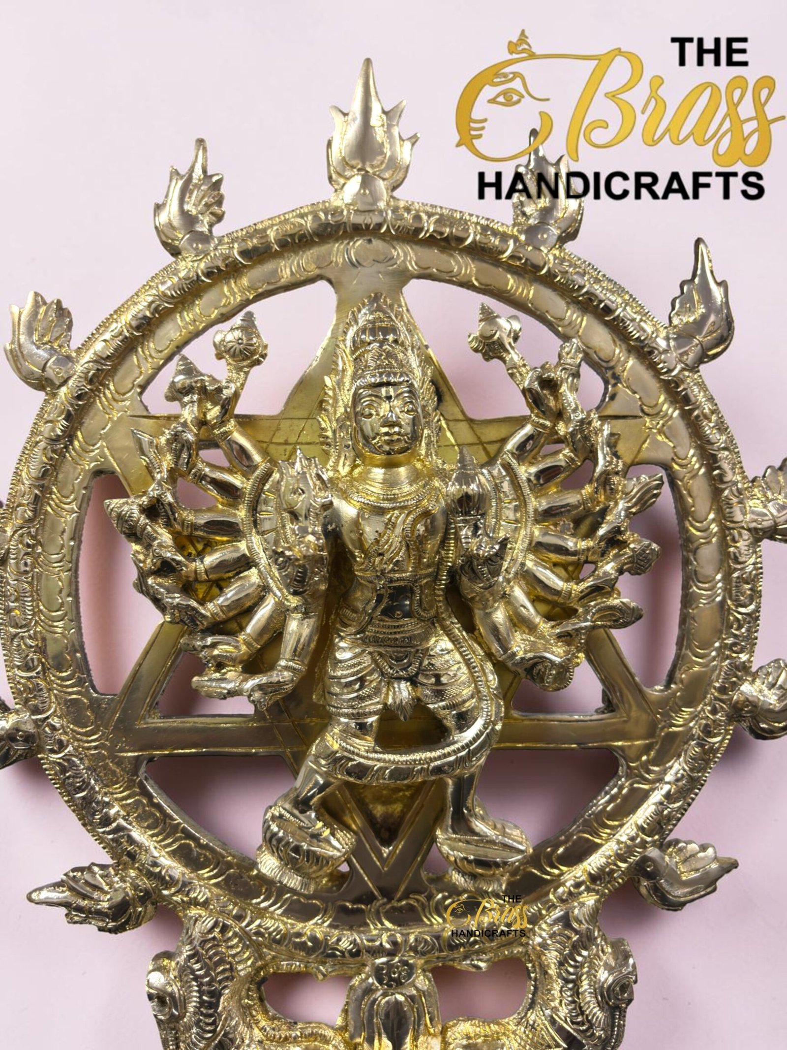 Gold brass statue of a multi-armed Hindu deity inside an ornate circular frame, on a pale pink background, with 'The Brass Handicrafts' logo visible.  thebrasshandicrafts.com