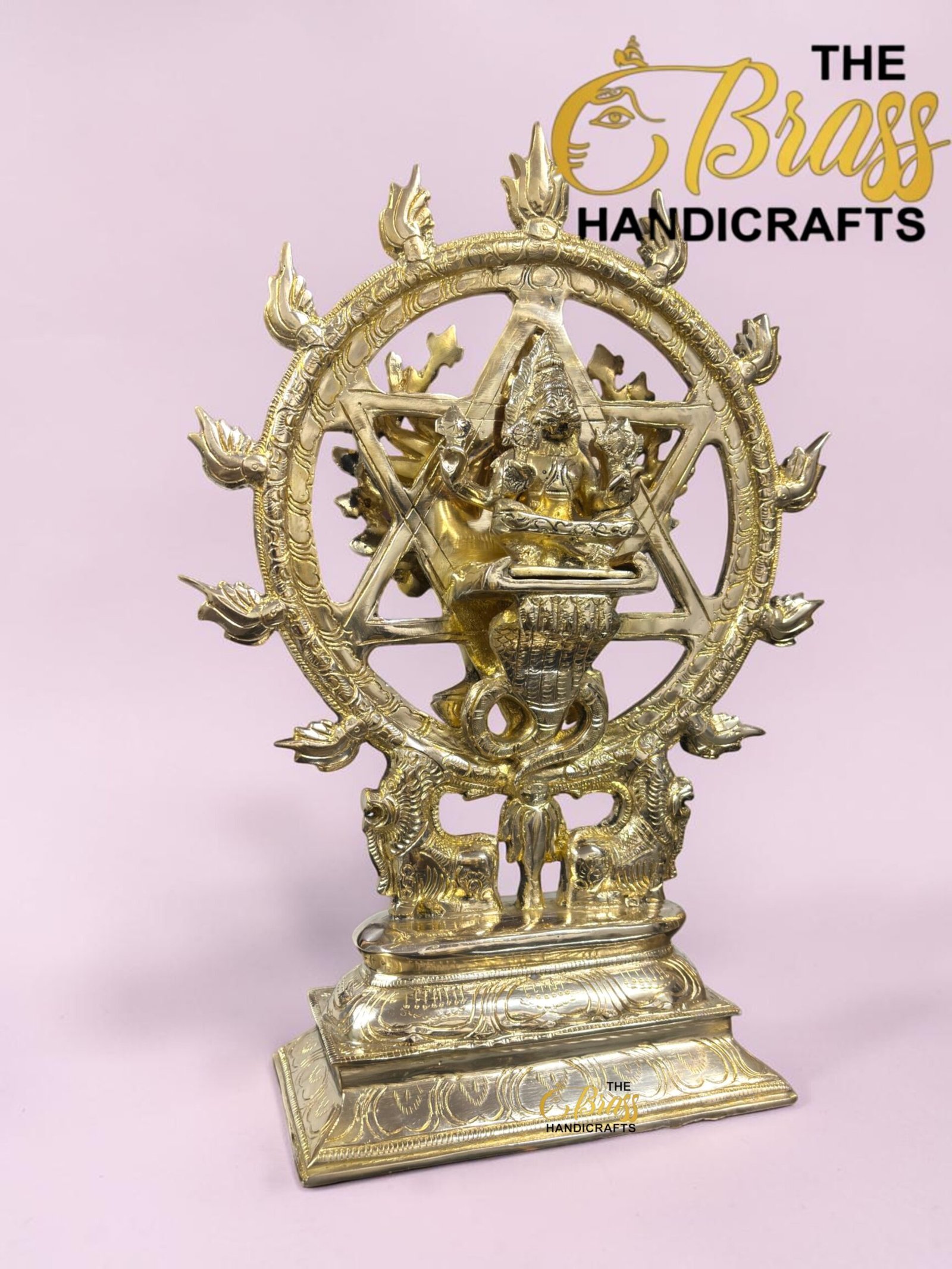 Ornate brass Nataraja statue (Shiva) dancing inside a circular ring, on a decorative pedestal with a pink background and 'The Brass Handicrafts' watermark.  thebrasshandicrafts.com