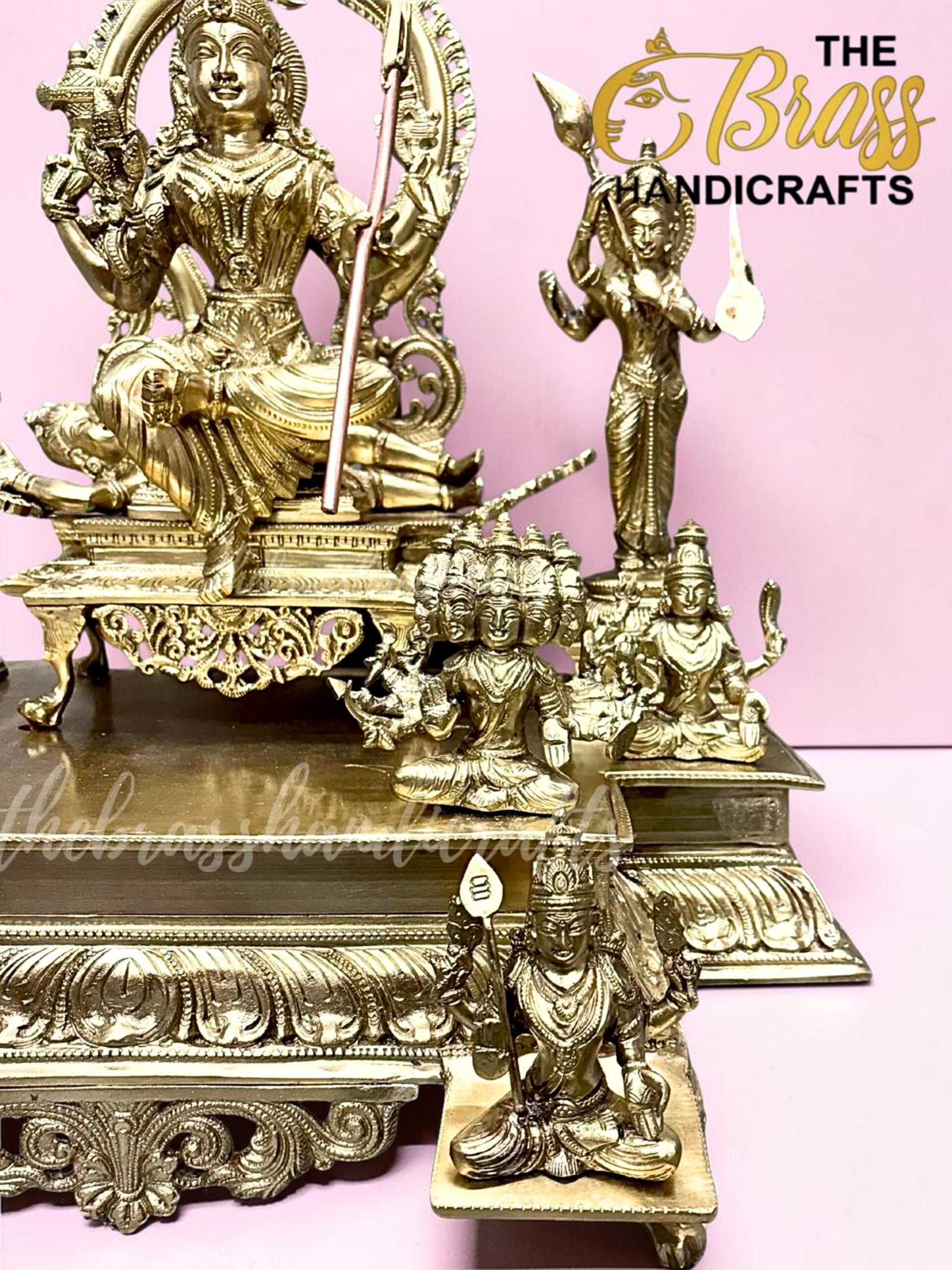 Intricately detailed gold-tone brass statues, with a central seated deity holding a staff and smaller figures around them against a pink backdrop.  thebrasshandicrafts.com