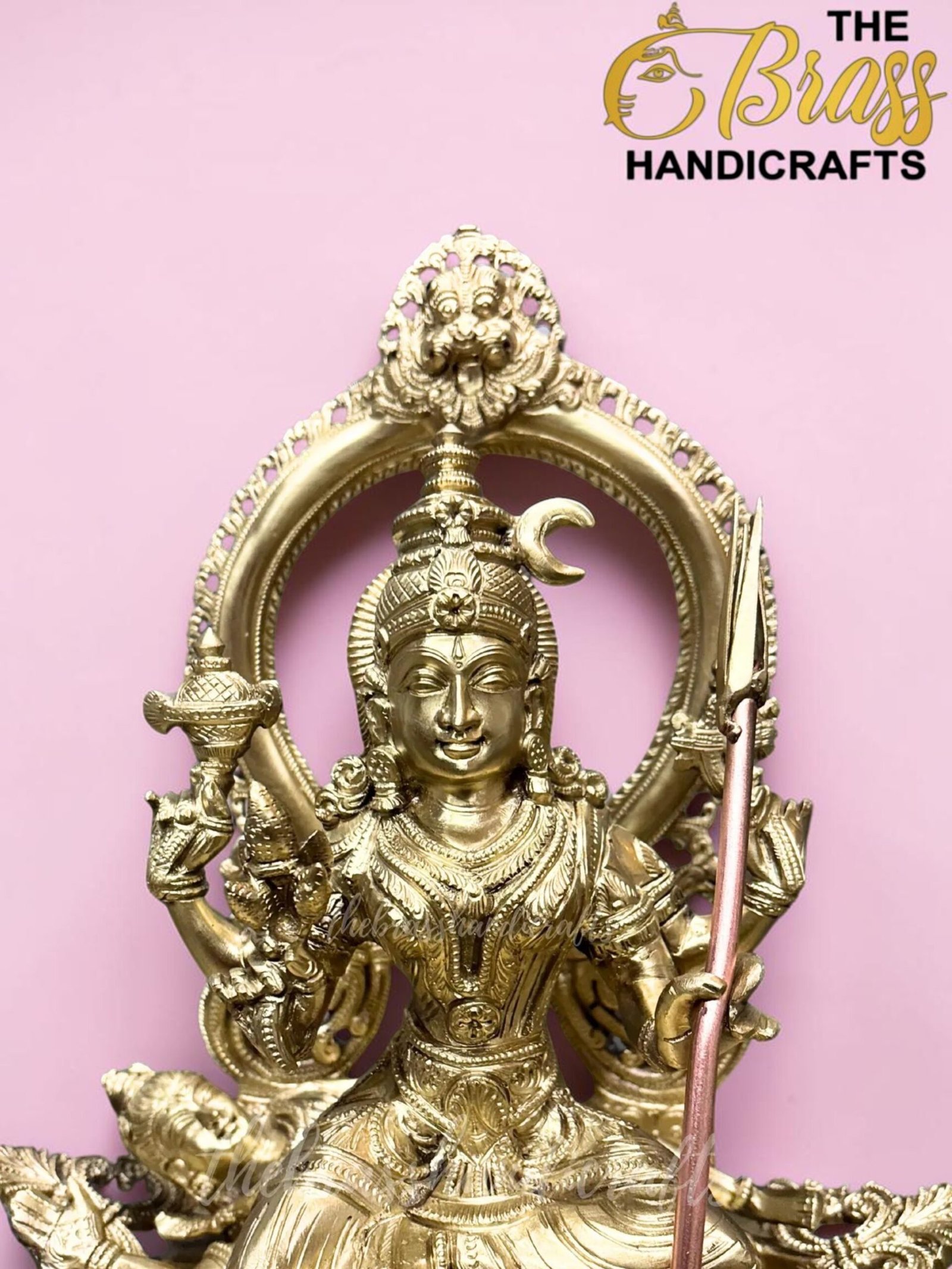 Ornate brass statue of a Hindu deity with a circular halo, set against a pink background.  thebrasshandicrafts.com