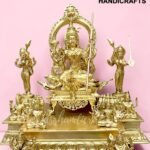Golden ornate brass statue of a seated deity with supporting figures on a decorative platform, pink backdrop, logo in corner.  thebrasshandicrafts.com