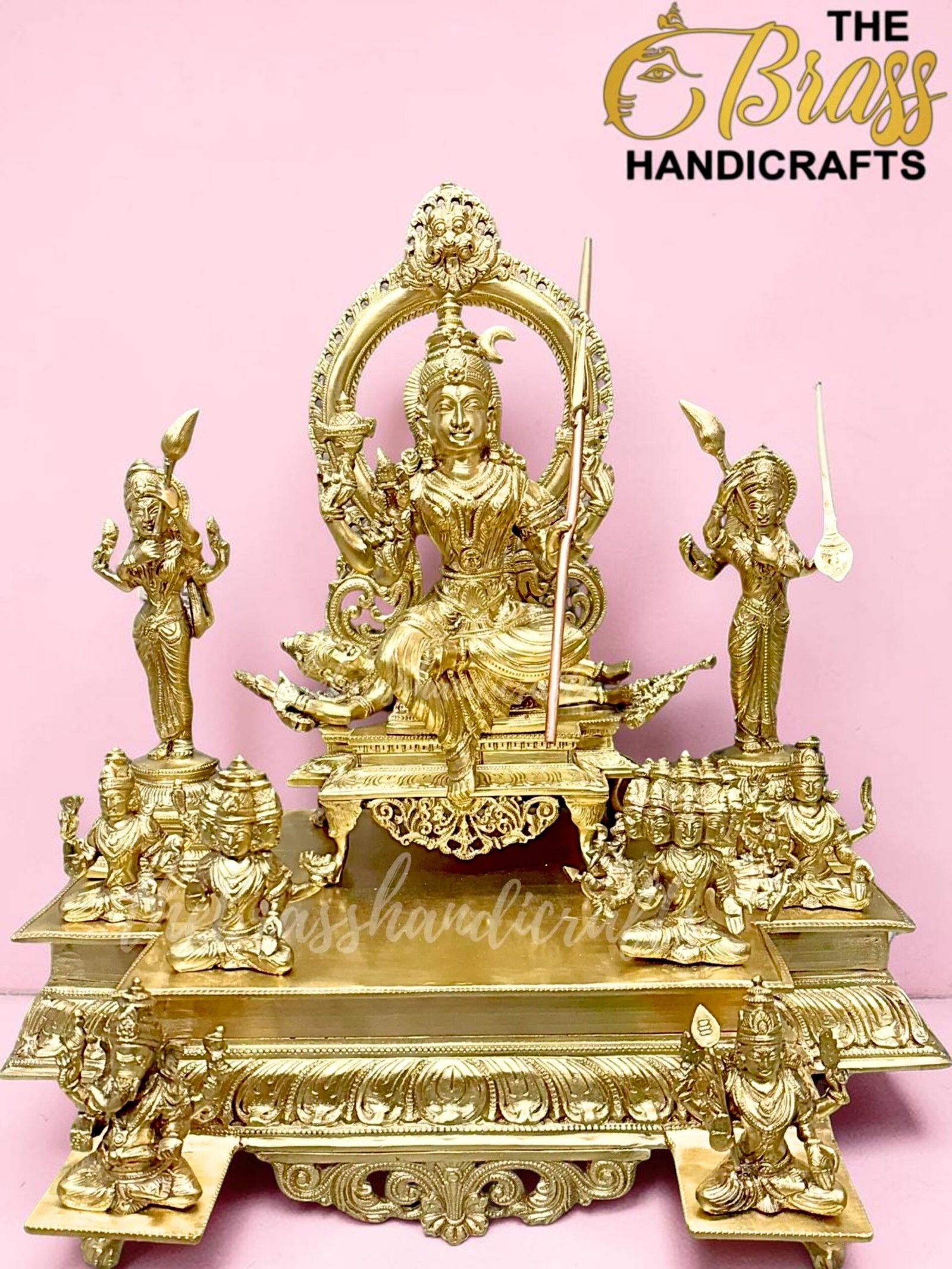 Golden ornate brass statue of a seated deity with supporting figures on a decorative platform, pink backdrop, logo in corner.  thebrasshandicrafts.com