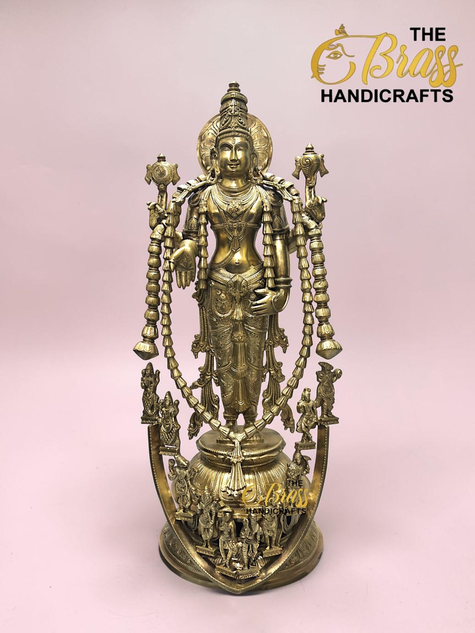Brass statue of a multi-armed Hindu deity with ornate jewelry and garlands, standing on a decorative base against a pink background.  thebrasshandicrafts.com