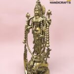 Brass statue of a Hindu goddess with an ornate headdress and multiple adornments, set against a pink background, with The Brass Handicrafts logo in the corner.  thebrasshandicrafts.com
