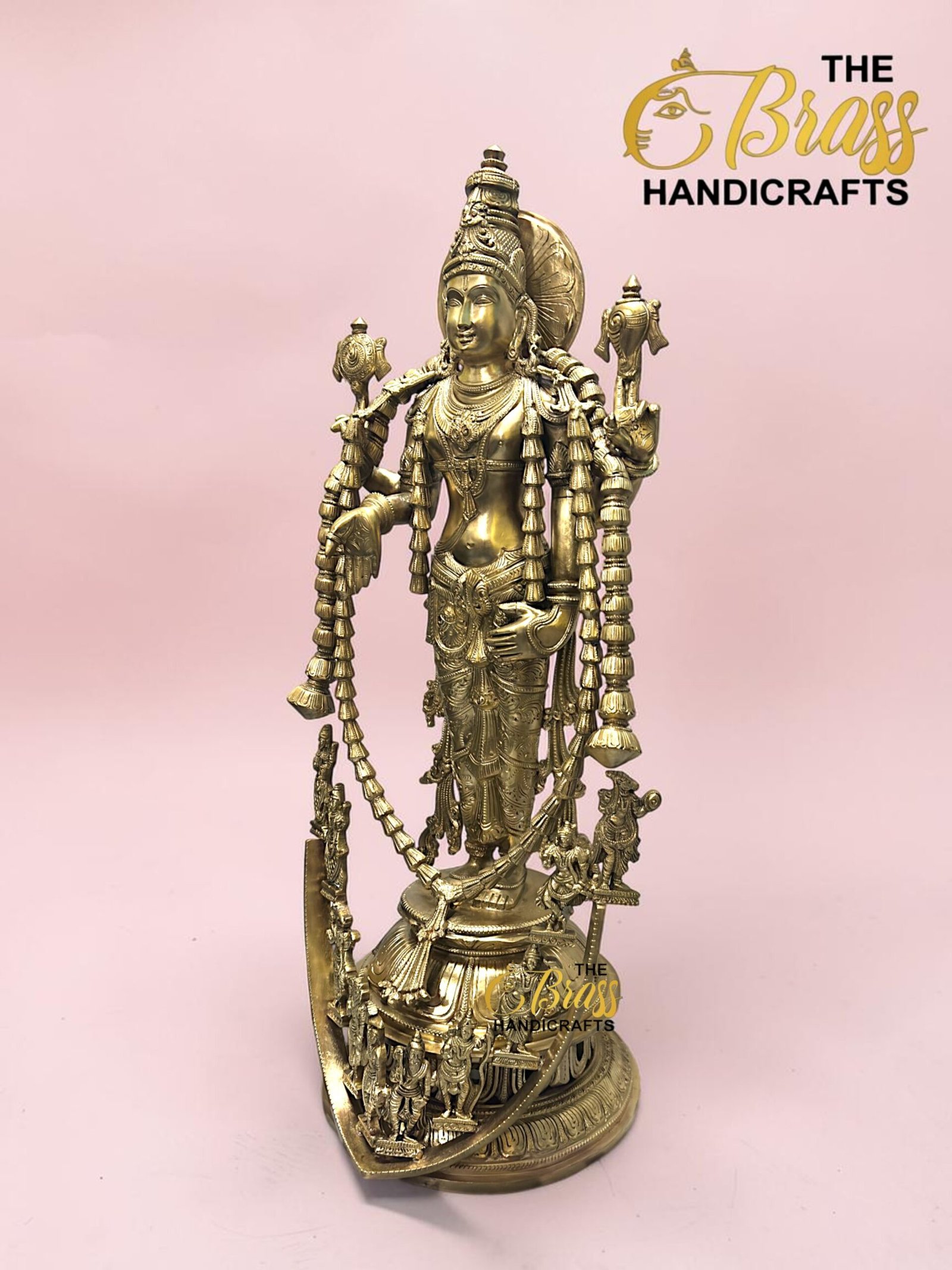 Brass statue of a Hindu goddess with an ornate headdress and multiple adornments, set against a pink background, with The Brass Handicrafts logo in the corner.  thebrasshandicrafts.com