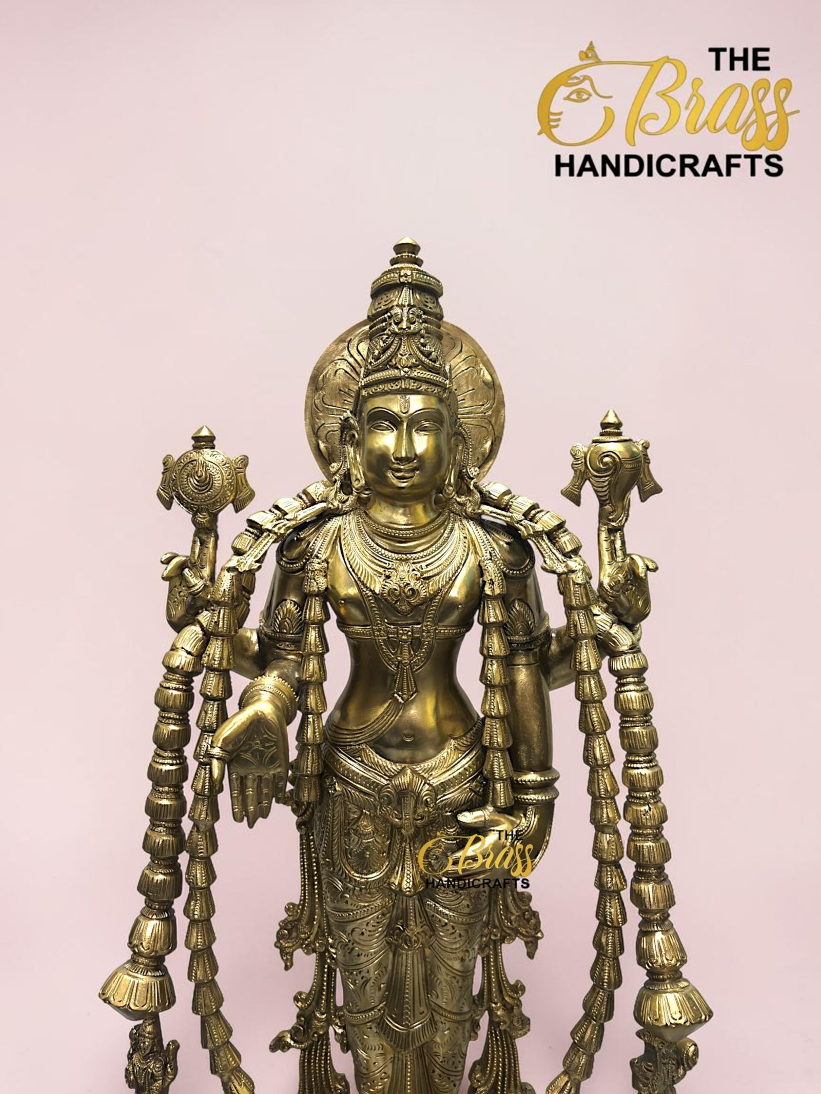 Gold brass statue of a multi-armed Hindu deity with ornate jewelry, set against a pale pink background; logo 'The Brass Handicrafts' appears in the top-right corner.  thebrasshandicrafts.com
