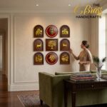 Wall with Indian-inspired gold plaque art featuring elephants and a central red round piece; a woman in beige touches a plaque on the right, with a peacock feather plant nearby.  thebrasshandicrafts.com