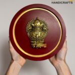 Round burgundy plaque with an ornate gold emblem being held, gold trim visible and a 'The Brass Handicrafts' watermark in the corner  thebrasshandicrafts.com