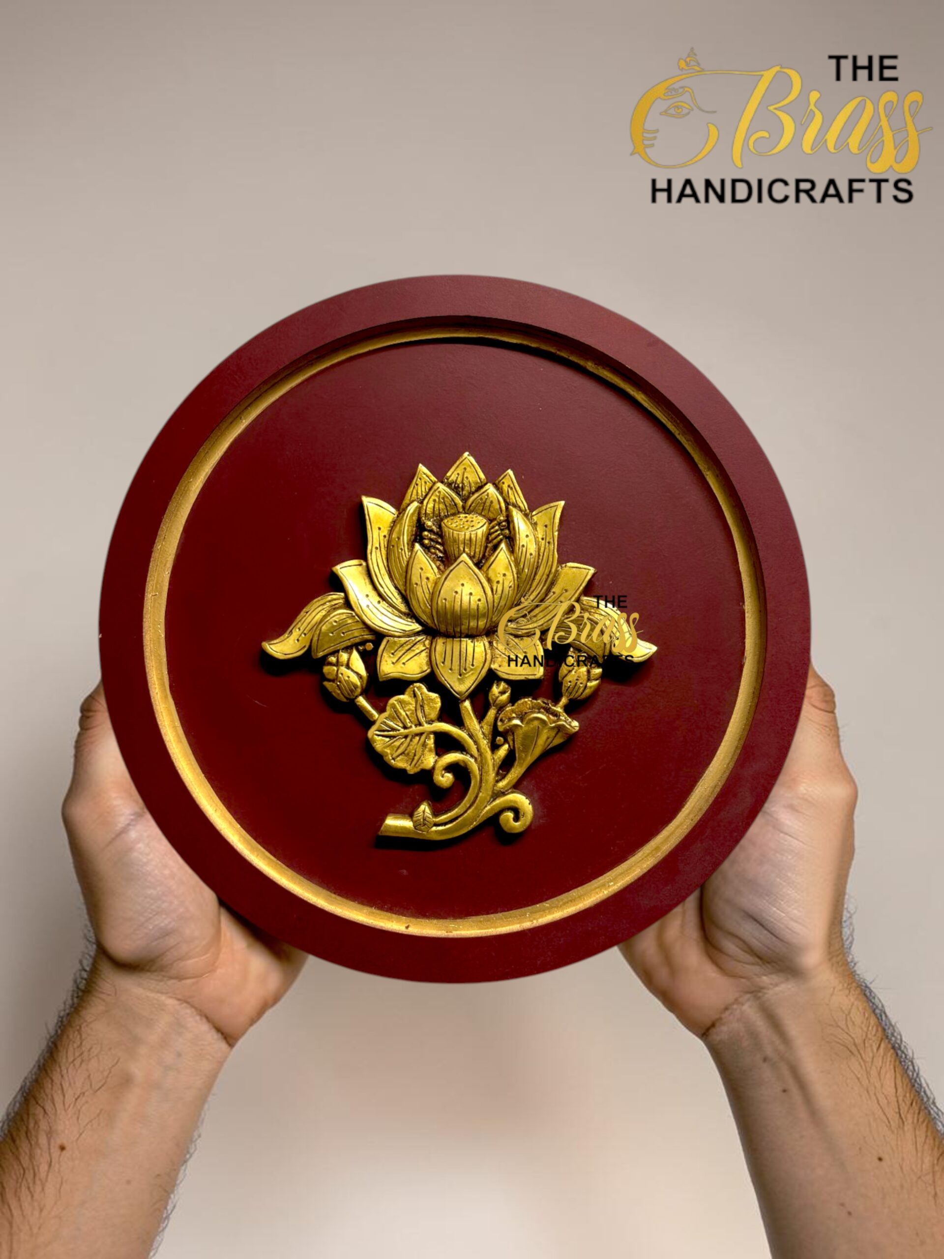 Red circular brass plaque with a gold lotus floral ornament being held by two hands; logo 'The Brass Handicrafts' appears in the upper corner.  thebrasshandicrafts.com