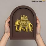 Gold decorative brass elephant statue mounted in a brown arched display, held by hands on both sides.  thebrasshandicrafts.com