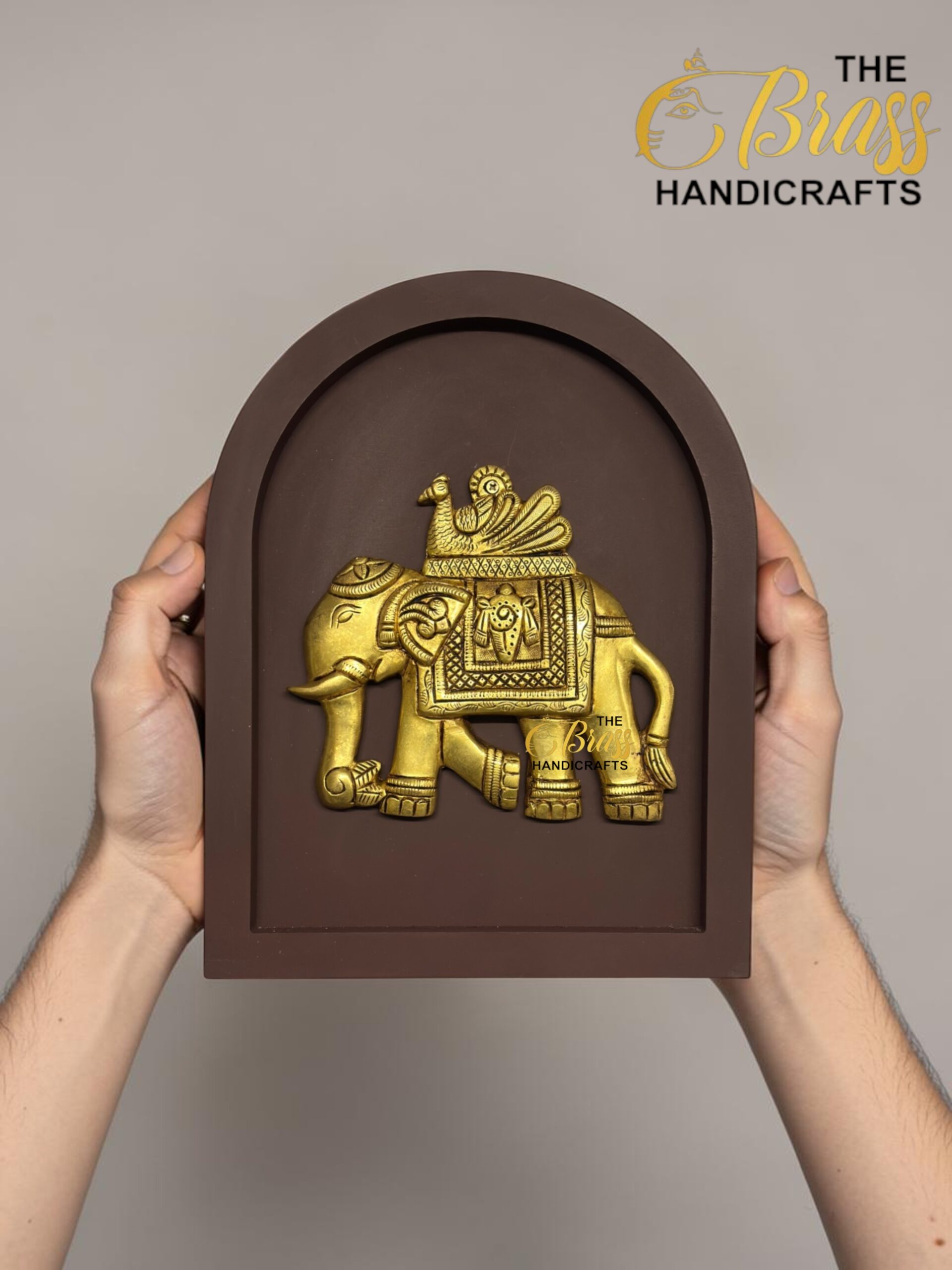 Gold decorative brass elephant statue mounted in a brown arched display, held by hands on both sides.  thebrasshandicrafts.com