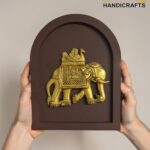 Gold-toned elephant statue mounted in a brown arched display frame, held by two hands against a neutral background; branding in corner reads 'The Brass Handicrafts'.  thebrasshandicrafts.com
