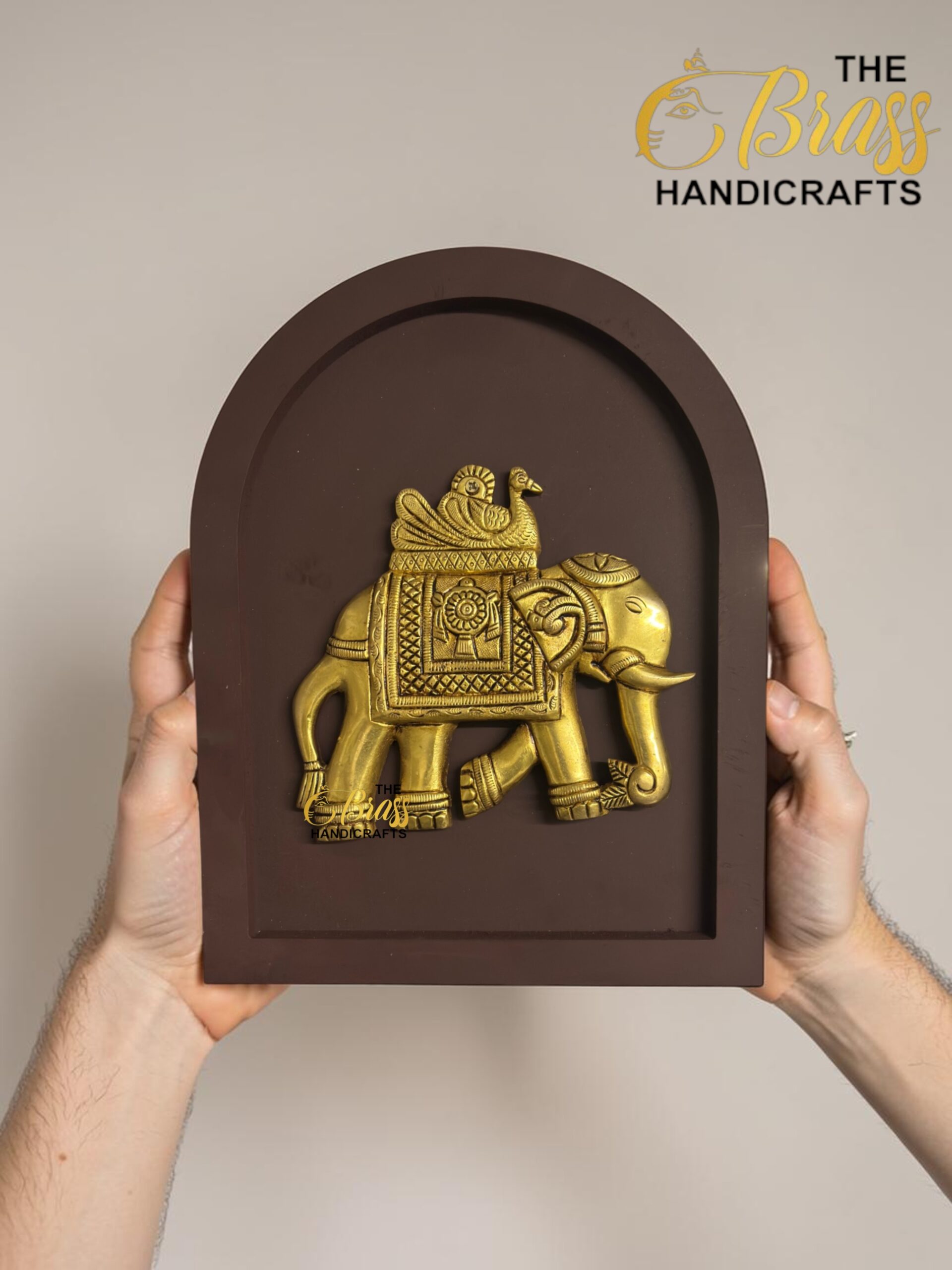 Gold-toned elephant statue mounted in a brown arched display frame, held by two hands against a neutral background; branding in corner reads 'The Brass Handicrafts'.  thebrasshandicrafts.com