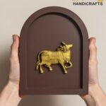 Brown arched plaque with a gold elephant sculpture held by two hands; logo 'The Brass Handicrafts' in top-right corner.  thebrasshandicrafts.com
