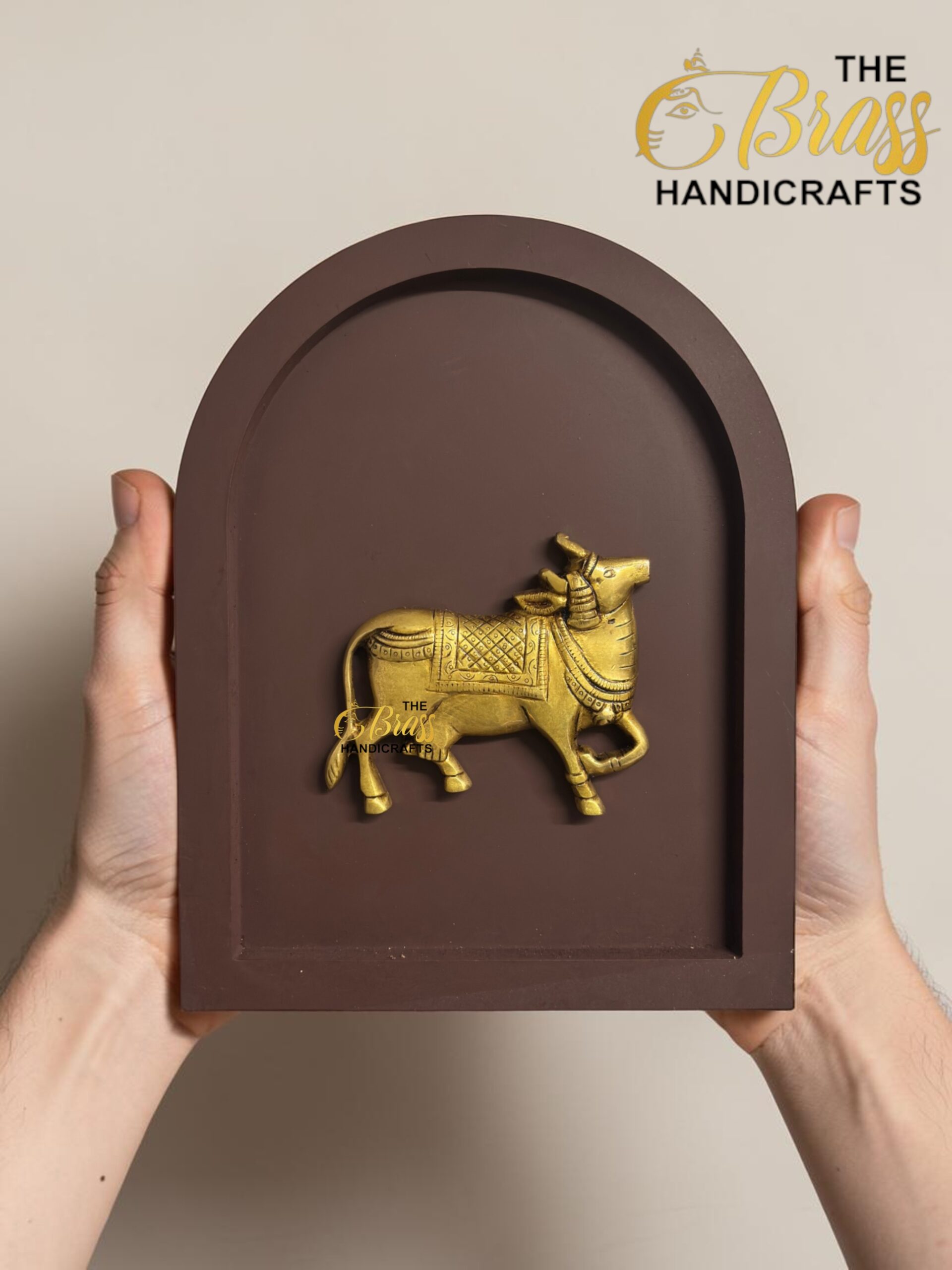 Brown arched plaque with a gold elephant sculpture held by two hands; logo 'The Brass Handicrafts' in top-right corner.  thebrasshandicrafts.com