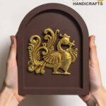 Brown arched decorative plaque with an intricate gold relief of a bird and scrolling foliage, held by hands against a light background.  thebrasshandicrafts.com