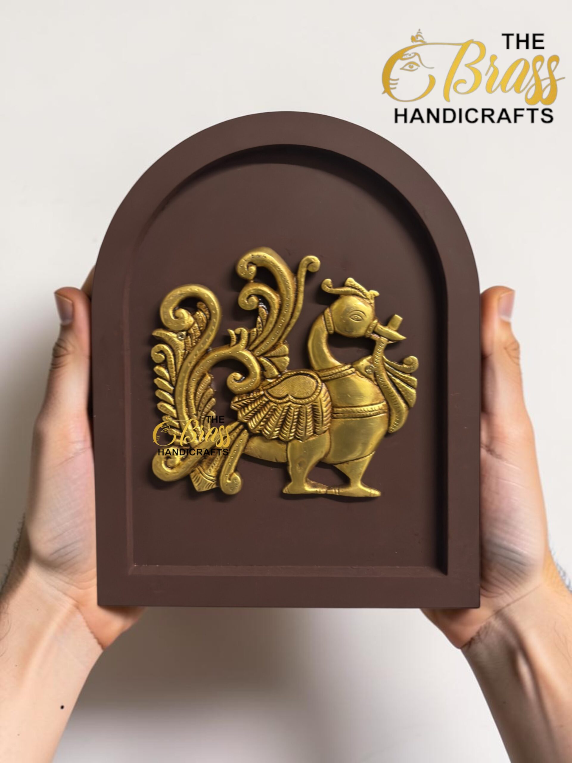 Brown arched decorative plaque with an intricate gold relief of a bird and scrolling foliage, held by hands against a light background.  thebrasshandicrafts.com