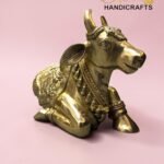 Gold-toned, ornately carved cow statue on a pink background with 'The Brass Handicrafts' logo in the top-right corner  thebrasshandicrafts.com