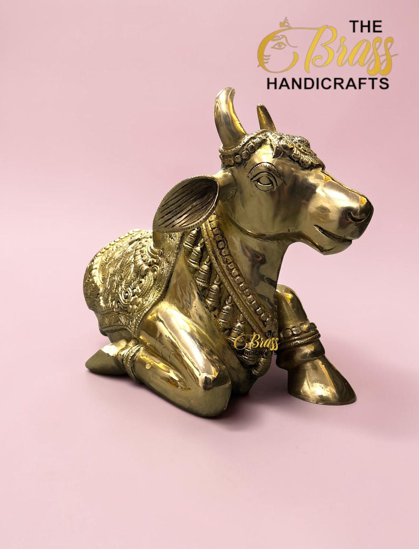 Gold-toned, ornately carved cow statue on a pink background with 'The Brass Handicrafts' logo in the top-right corner  thebrasshandicrafts.com