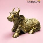 Gold-toned brass cow statue with intricate engravings on a pink background, featuring 'The Brass Handicrafts' logo in the corner.  thebrasshandicrafts.com