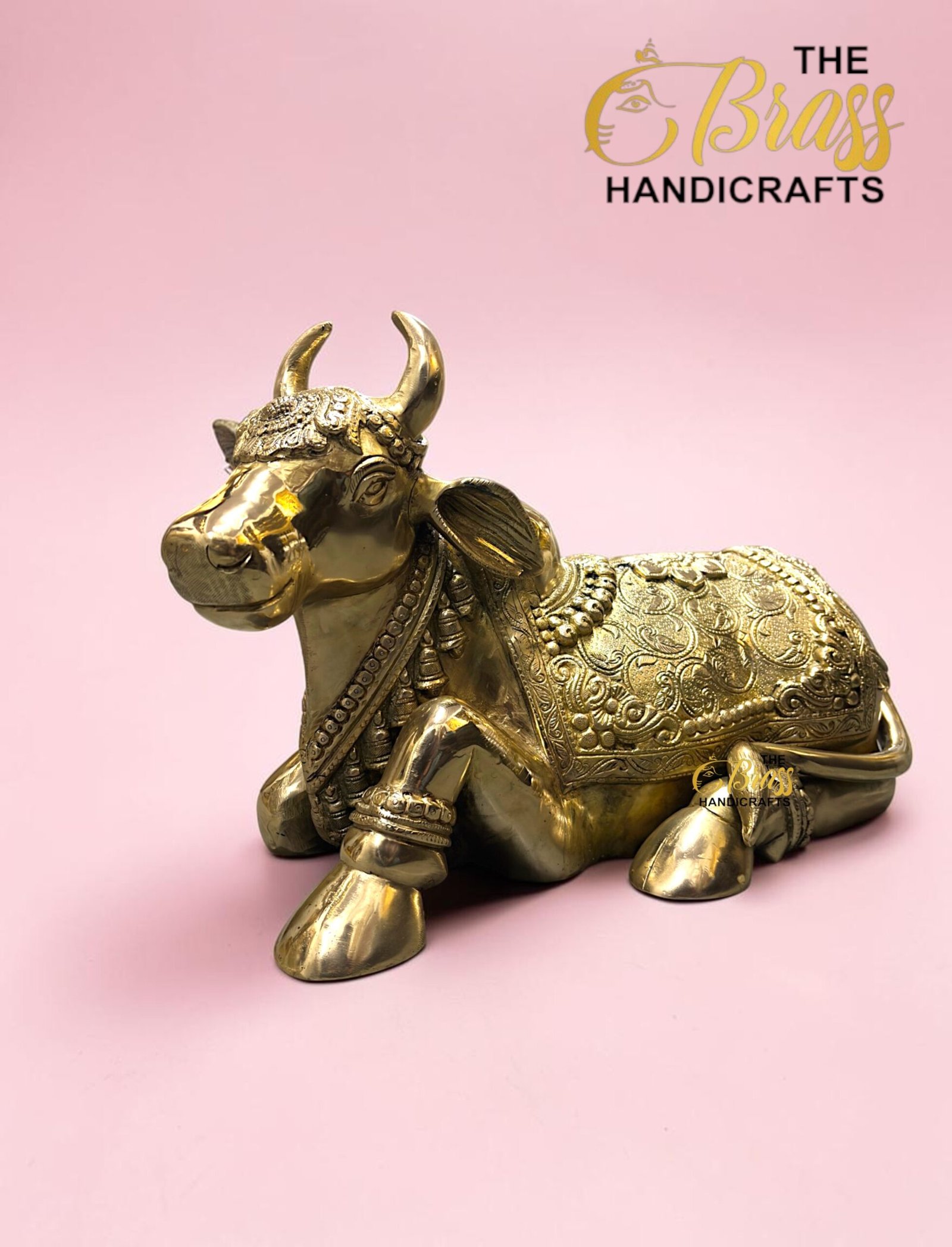 Gold-toned brass cow statue with intricate engravings on a pink background, featuring 'The Brass Handicrafts' logo in the corner.  thebrasshandicrafts.com