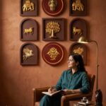 Woman sits in a leather chair writing in a notebook beside a wooden table, wall art with gold accents behind her.  thebrasshandicrafts.com