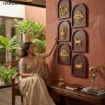 Sampada 1 - thebrasshandicrafts.com A woman in a beige saree sits and touches decorative wall plaques with gold motifs in a showroom, logo reading The Brass Handicrafts in the corner. thebrasshandicrafts.com