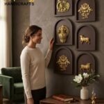 sampada - thebrasshandicrafts.com Woman in a beige sweater admires gold decorative reliefs mounted on a brown wall, with 'The Brass Handicrafts' logo in the top left corner. thebrasshandicrafts.com