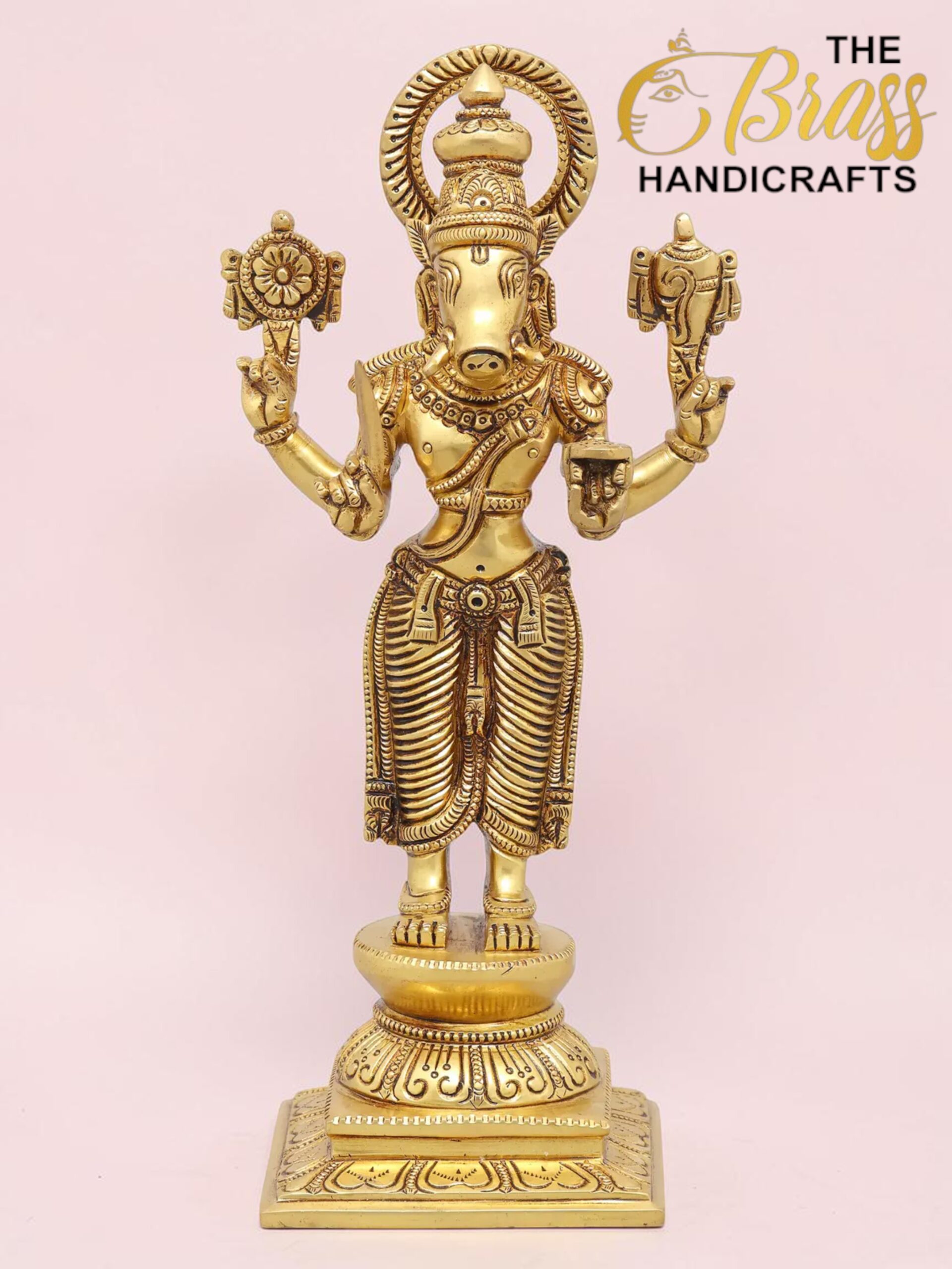 Gold-toned brass statue of the Hindu god Ganesha with an elephant head and multiple arms, holding symbolic objects, on a decorative base against a pink backdrop.  thebrasshandicrafts.com