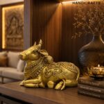 nandi 1 - thebrasshandicrafts.com Gold brass cow statue with ornate patterns on a wooden shelf, beside a dark vase and stacked books. thebrasshandicrafts.com