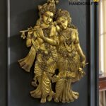 Golden statue of Krishna playing a flute with Radha beside him, mounted on a dark wall; watermark reads The Brass Handicrafts.  thebrasshandicrafts.com