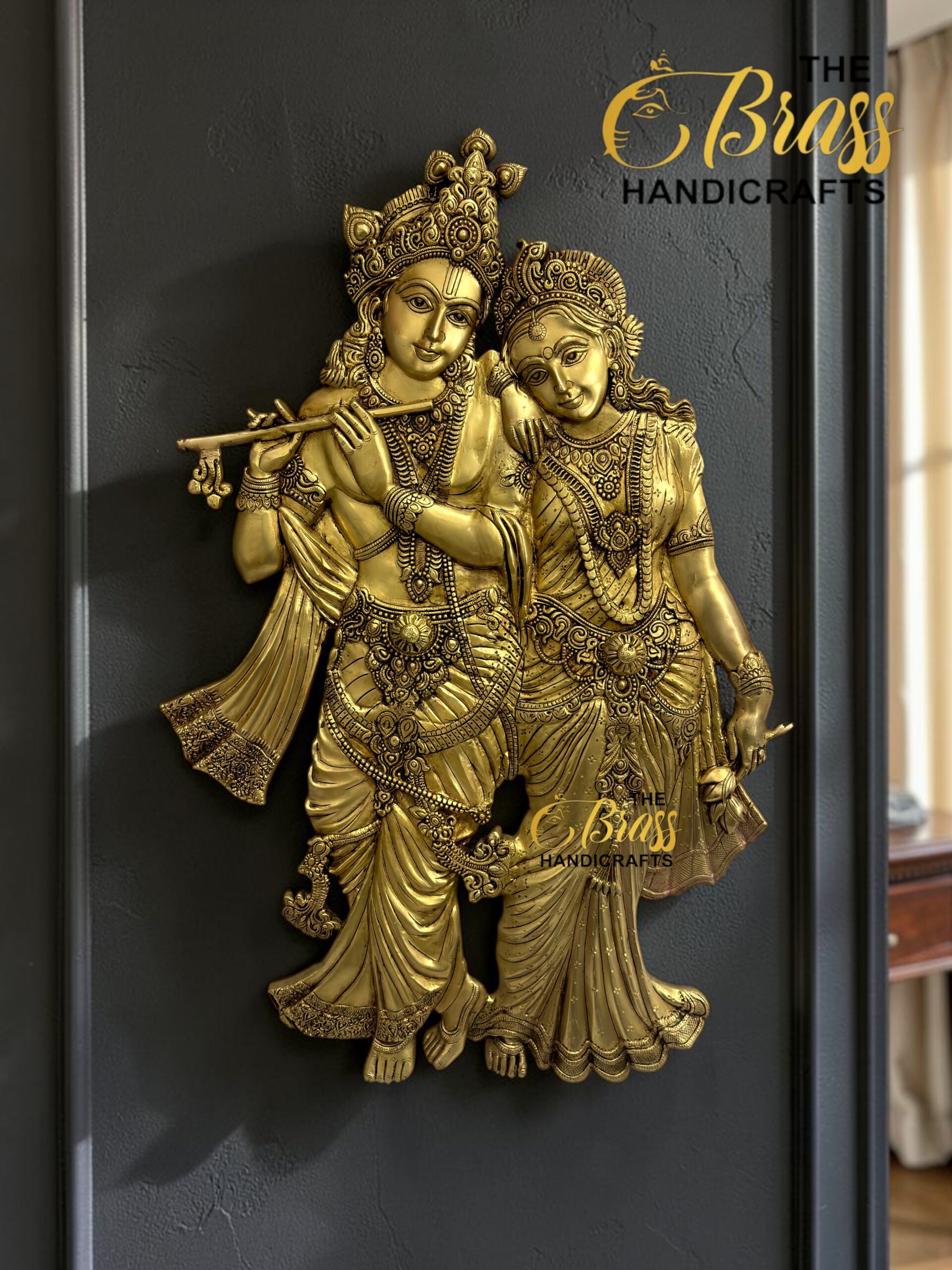 Golden statue of Krishna playing a flute with Radha beside him, mounted on a dark wall; watermark reads The Brass Handicrafts.  thebrasshandicrafts.com