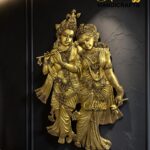 Gold-toned statue of Krishna playing a flute with Radha beside him against a dark textured wall.  thebrasshandicrafts.com