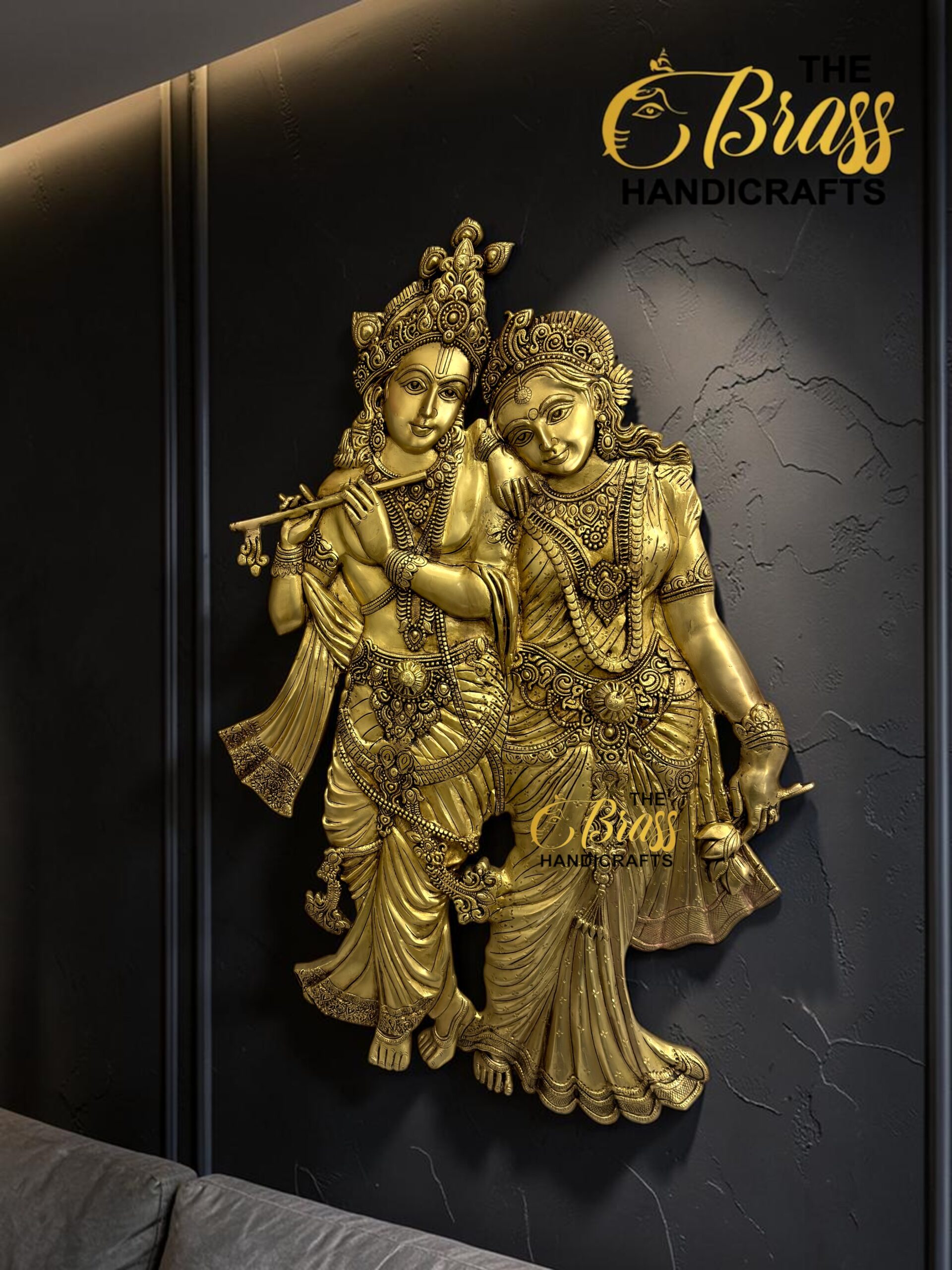 Gold-toned statue of Krishna playing a flute with Radha beside him against a dark textured wall.  thebrasshandicrafts.com