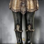 Lower half of an ornate black-and-bronze statue with detailed armor and sandals, standing on a round base; a logo reading 'The Brass Handicrafts' appears on the image.  thebrasshandicrafts.com