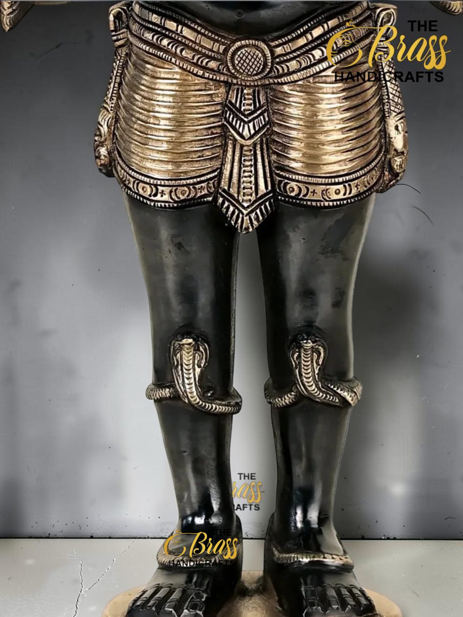 Lower half of an ornate black-and-bronze statue with detailed armor and sandals, standing on a round base; a logo reading 'The Brass Handicrafts' appears on the image.  thebrasshandicrafts.com