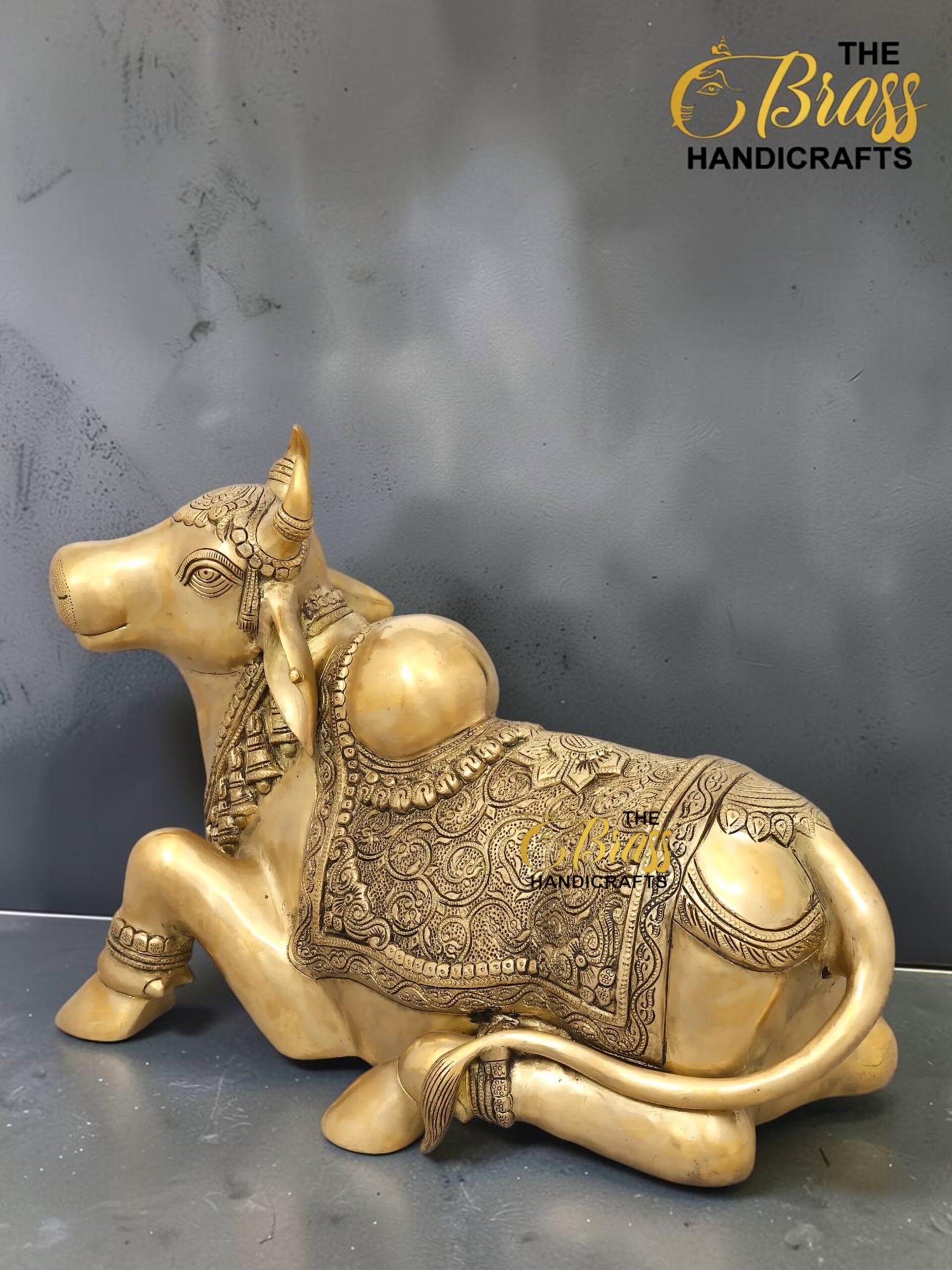 nandi 1 - thebrasshandicrafts.com Golden ornate reclining cow statue with intricate engravings and jewelry patterns, on a dark backdrop (The Brass Handicrafts watermark). thebrasshandicrafts.com